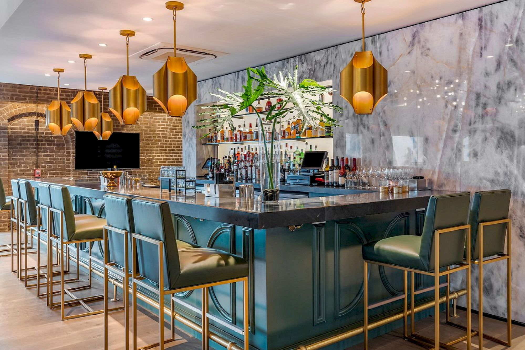 A stylish bar with a marble wall, gold light fixtures, green barstools, and a plant on the counter.