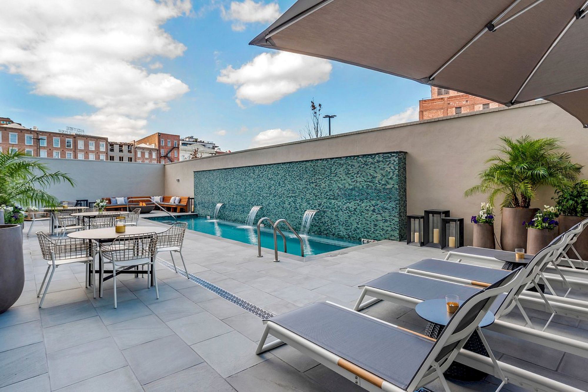 A rooftop pool with a waterfall feature, lounge chairs, and tables under a blue sky with clouds.